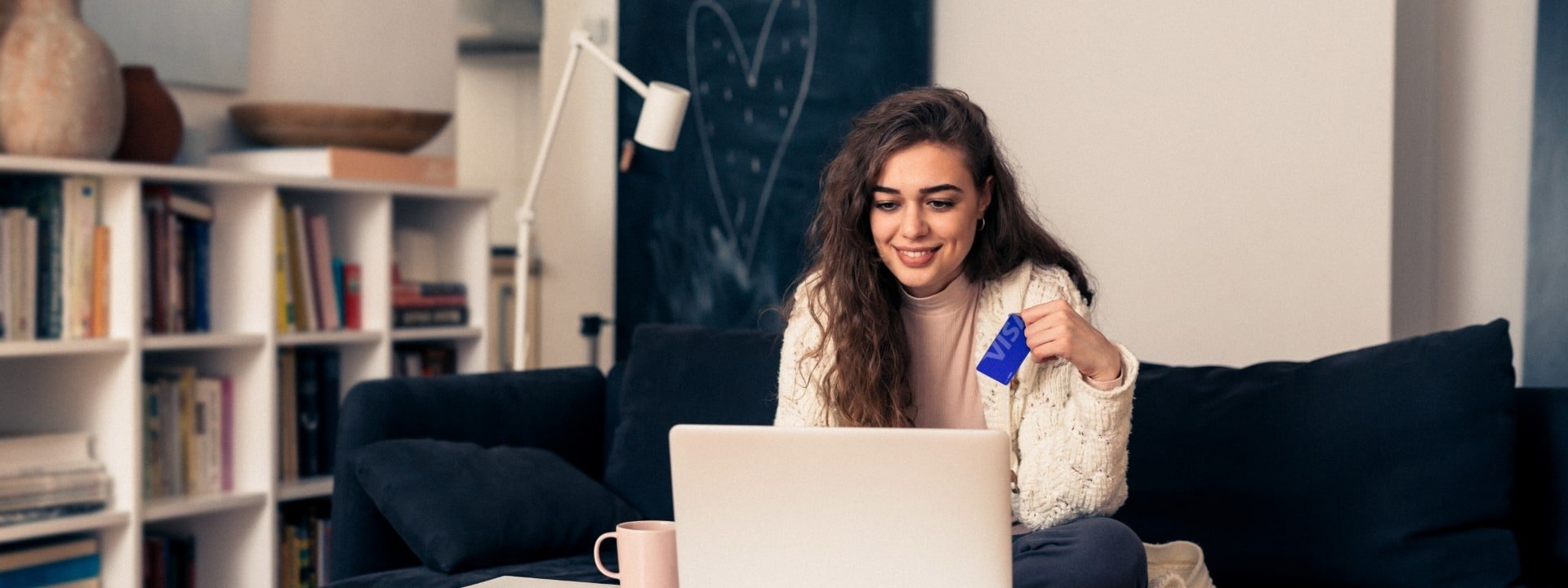 Young woman at home, smiling while using a laptop and holding a blue Visa card