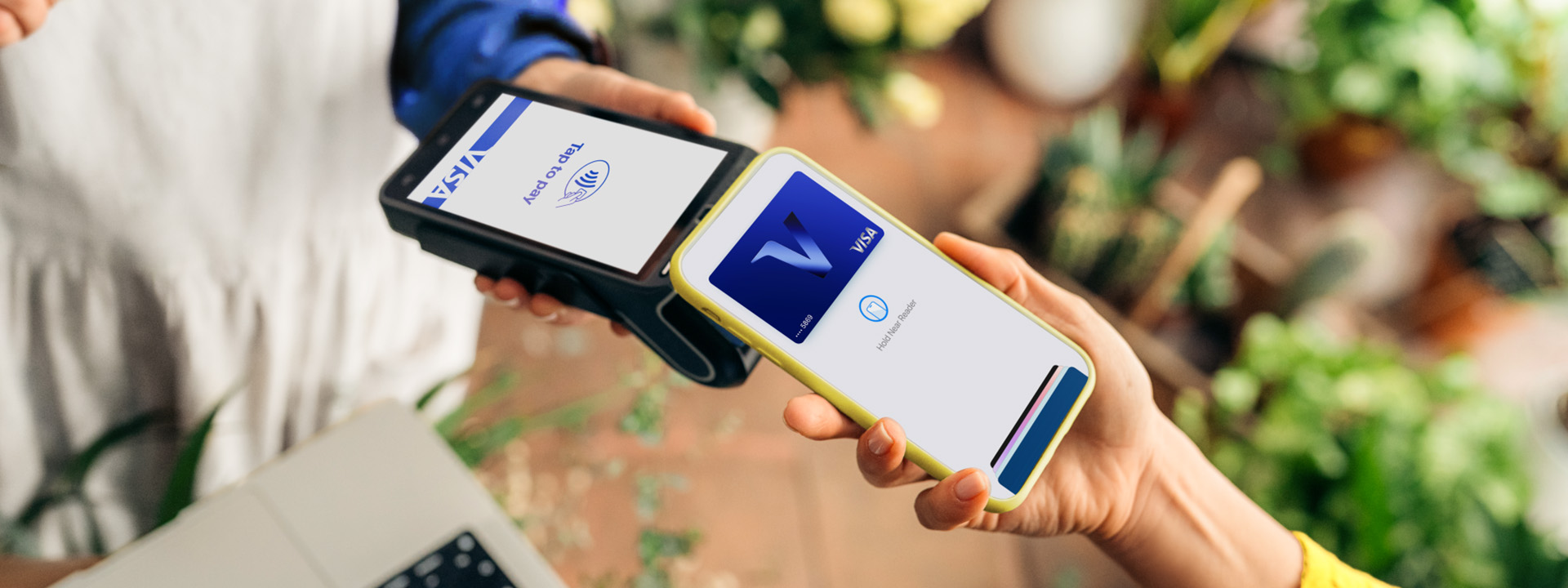 Customer holds a smartphone near a contactless payment terminal displaying Visa branding in a plant-filled shop.