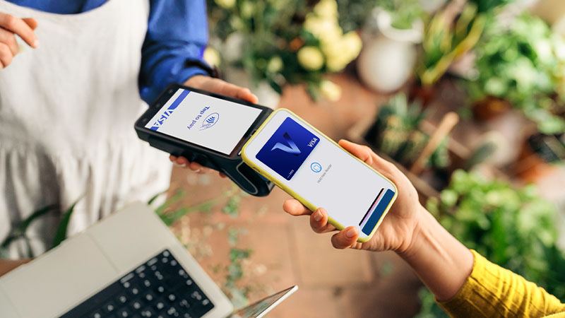 Customer holds a smartphone near a contactless payment terminal displaying Visa branding in a plant-filled shop.