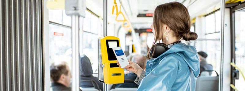 Woman in a blue slicker tapping her mobile wallet at a fare reader on a bus to pay her public transit fare