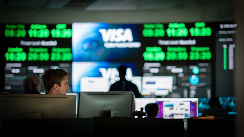 Visa employee working behind two computers in front of a wall of screens.