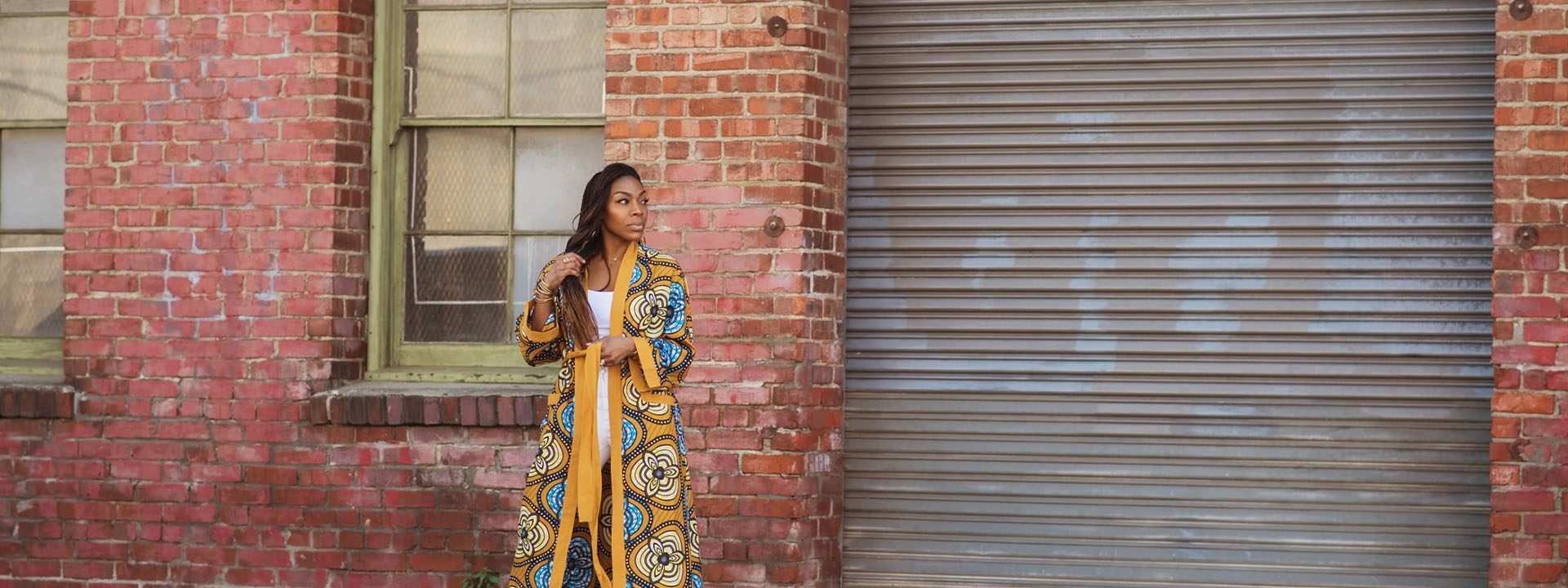 Love Iguehi founder Iguehi James wears her ankle-length Fula Duster in front of a closed corrugated steel garage door.