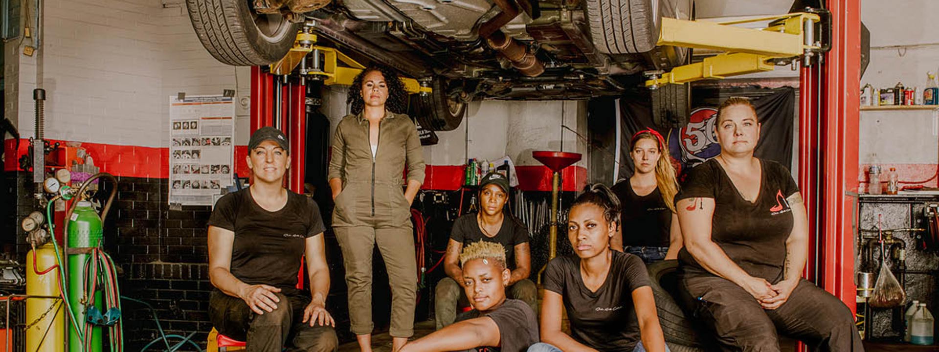 Seven women pose under an elevated car in the garage of Girls Auto Clinic.