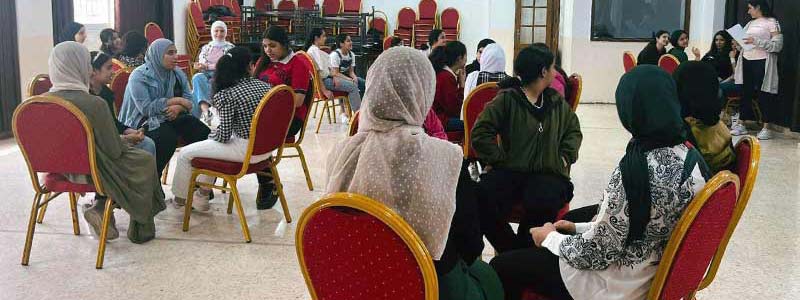 Girls and women sit around tables in a room listening to a speaker.