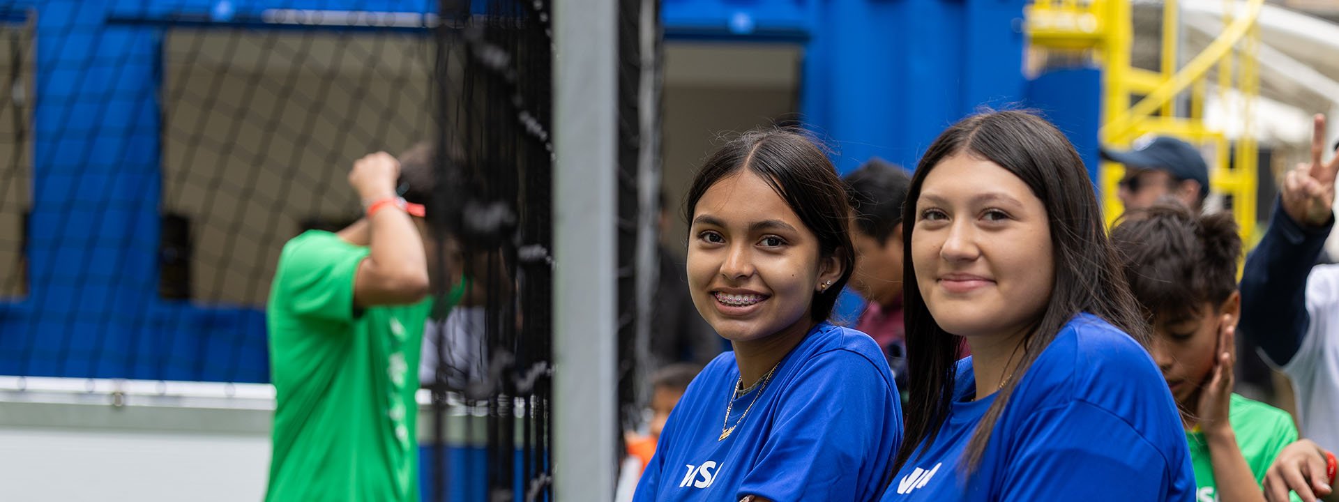 Two young soccer players wearing blue shirts stand next to the street soccer field, looking at the camera.