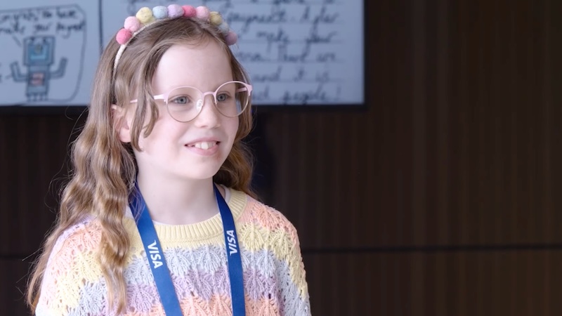 A young girl smiling wearing pink glasses and a Visa lanyard around her neck
