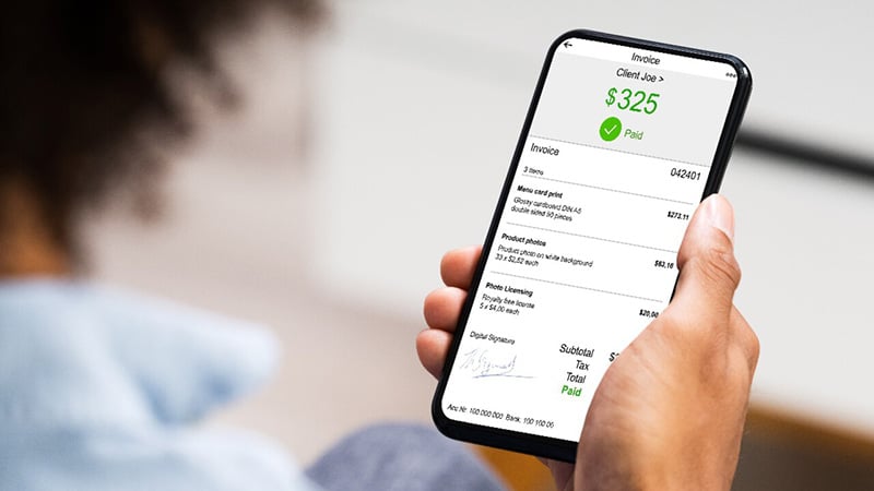 Right hand holding iPhone displaying "invoice" and "$325 paid" for menu card print, product photo, and photo licensing. The person's head is blurred.