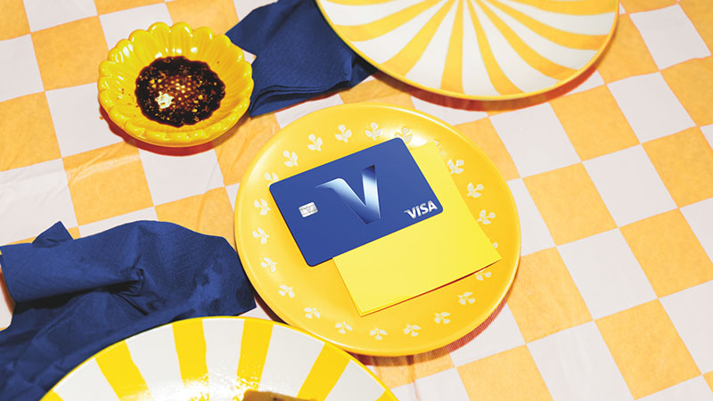 Blue Visa card displayed on a yellow plate atop a yellow-and-white checkered table with colorful patterned dishes.