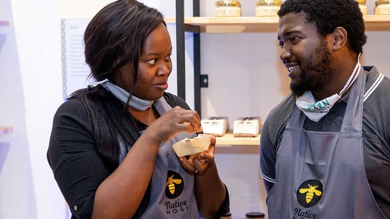 Two Native Nosi team members in branded aprons share a light moment while sampling honey in front of product shelves.