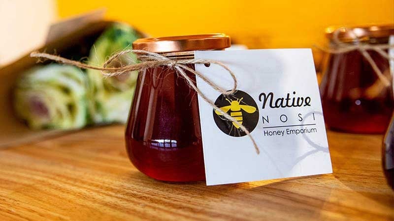 A small jar of honey with a copper lid sits on wood, tagged with the Native Nosi Honey Emporium logo and tied with twine.