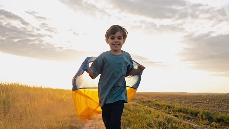 Boy running through field holding the Ukraine flag behind him.