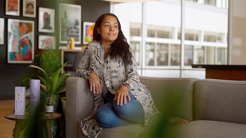 Woman sits on a couch in a room slightly smiling.