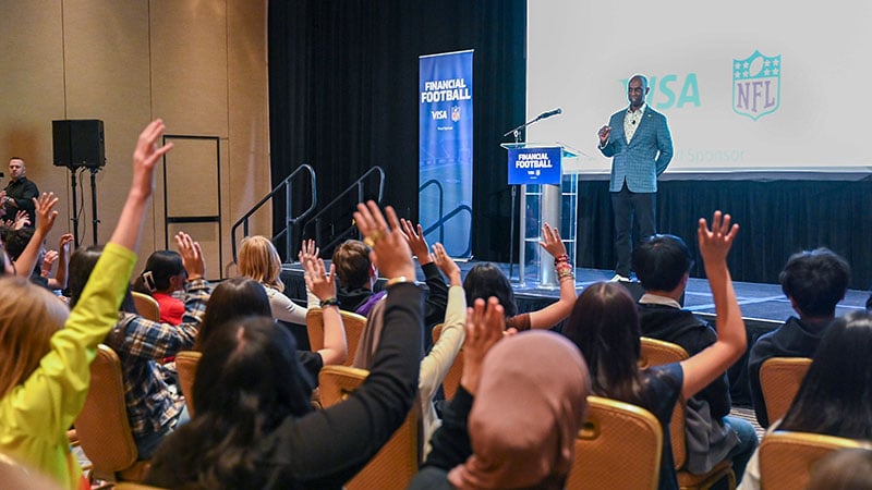 A room full of people raising their hands facing someone speaking on stage