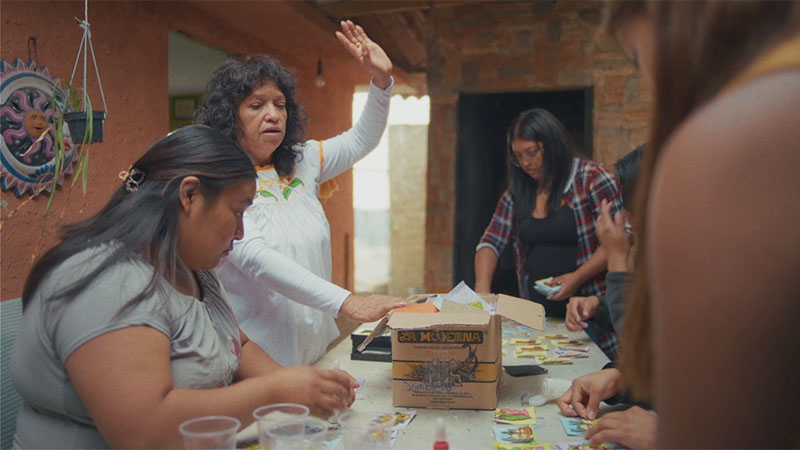 Five women standing and sitting around a table creating popotillo art.
