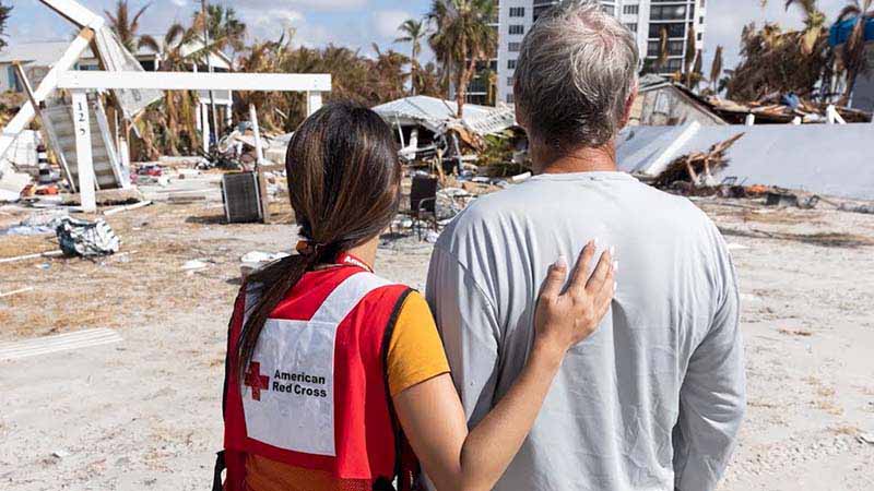 Woman wearing an American Red Cross vest stands next to a man looking out on a natural disaster site.