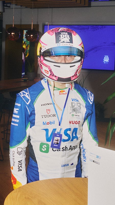 One driver wearing his racing gear and helmet stands behind an office desk with an ID badge around his neck.