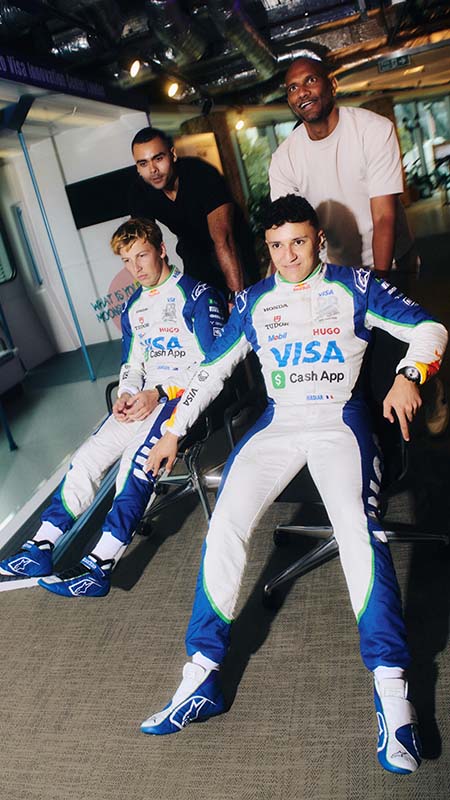 Two drivers wearing their racing gear sit in office chairs with two people behind them ready to push.