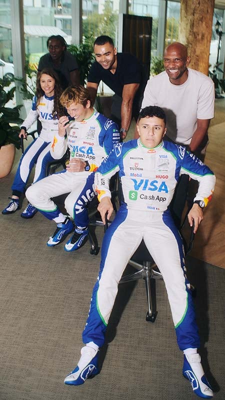 Three drivers wearing their racing gear sit in office chairs in an office hallway waiting to be pushed by the people behind them.