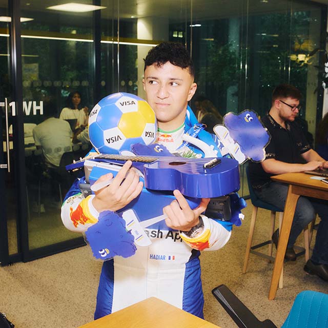 Driver Isack Hadjar standing in an office building holding a pile of Visa-branded items.